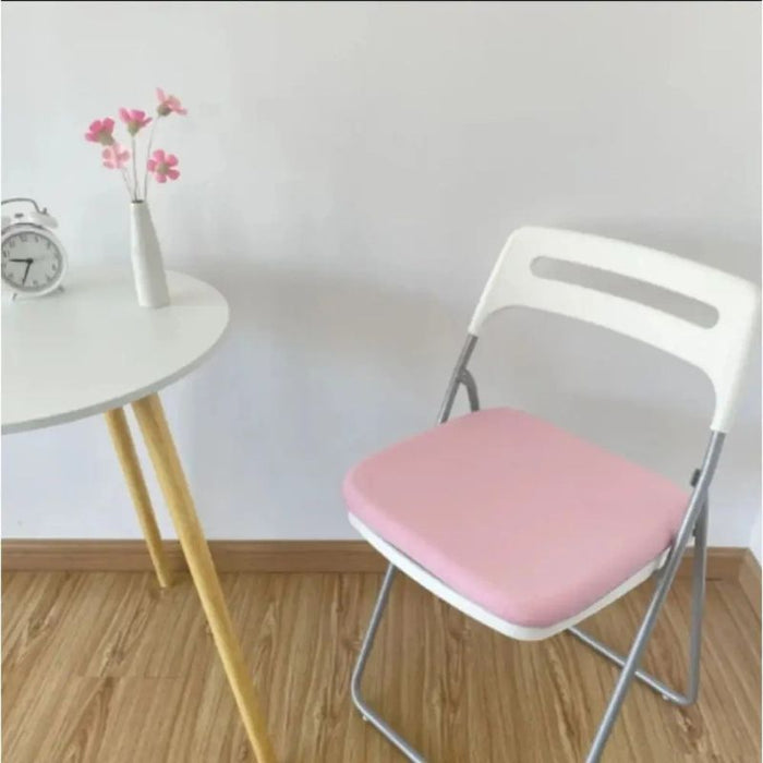 Cadeira dobrável branca com almofada ergonômica rosa sobre assento, ao lado de uma mesa branca e decoração minimalista.