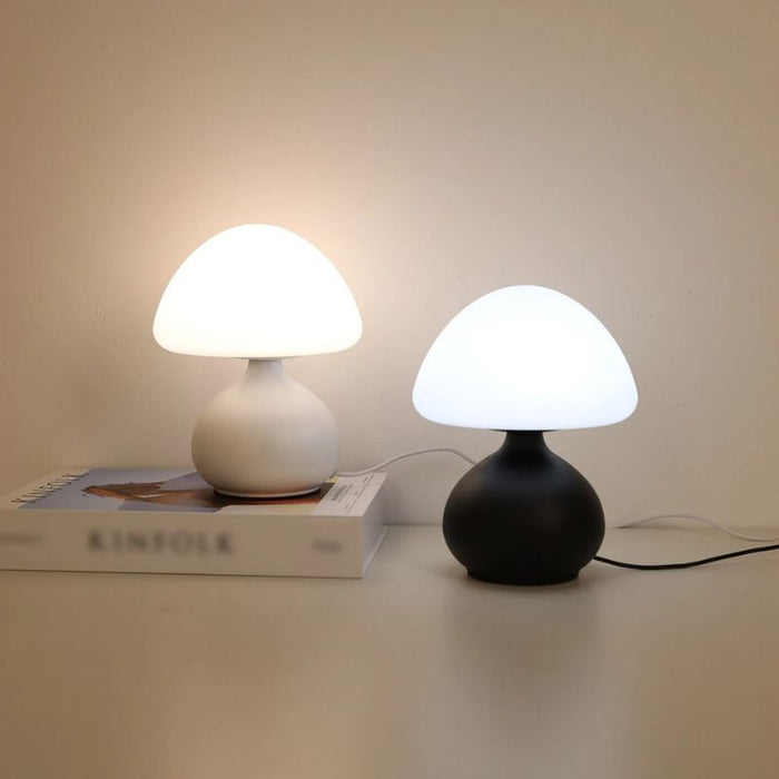 Duas luminárias de mesa estilo cogumelo, uma branca e uma preta, com luzes LED acesas sobre um livro decorativo.