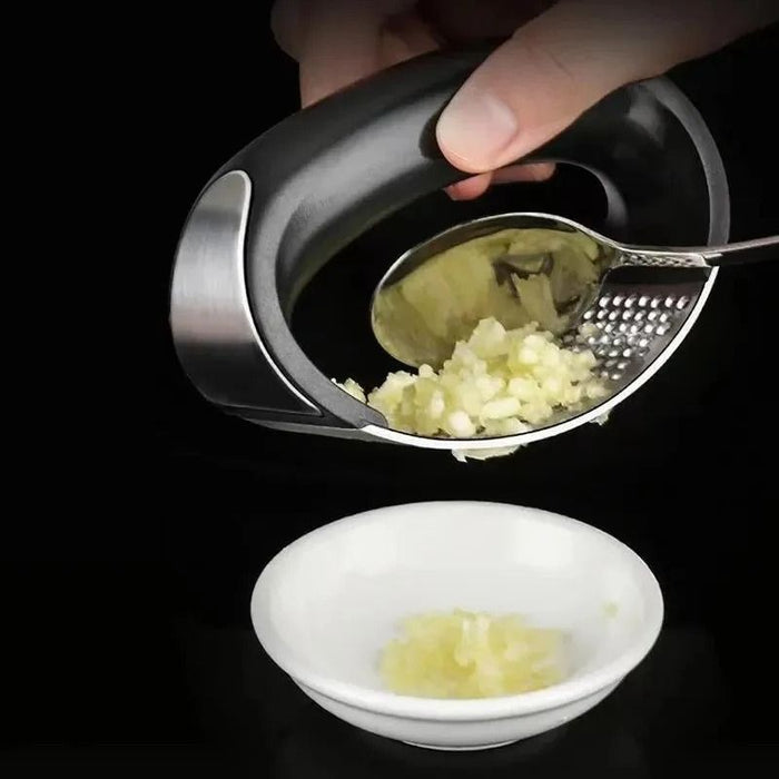 Mão utilizando espremedor de alho ergonômico de inox para triturar alho em tigela branca sobre fundo preto.